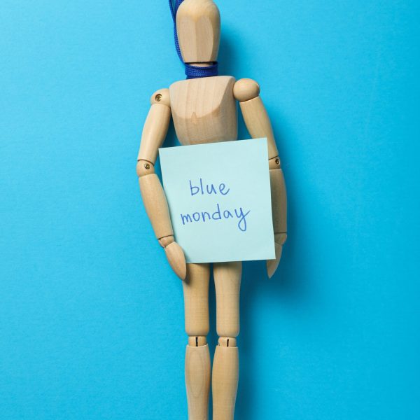 Wooden man on blue ribbon with paper with the text Blue Monday on blue background, top view
